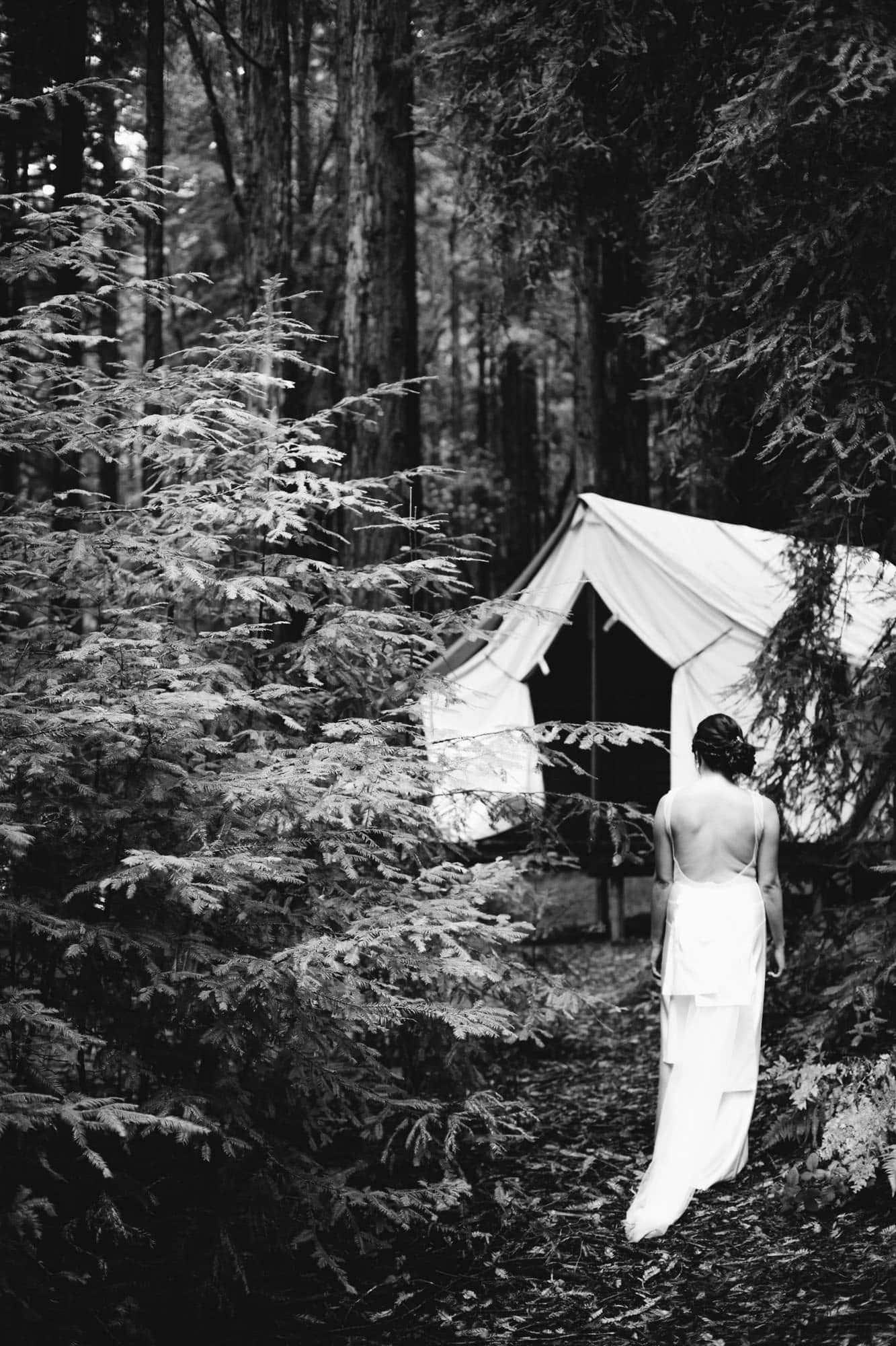 Black and white photo of the back of a brides dress in the woods in front of a tent