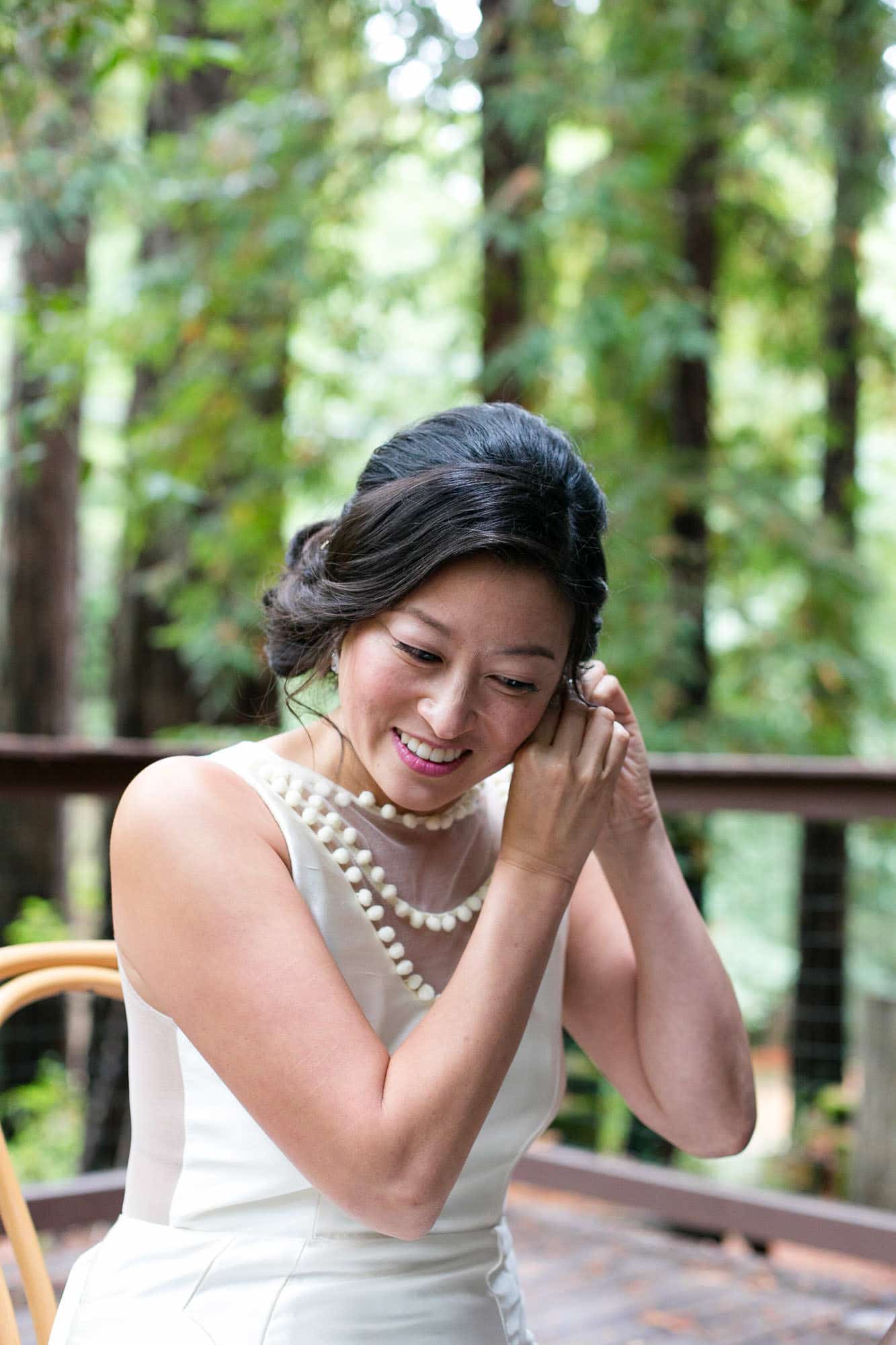 Bride in white dress sitting down outside while putting on her earrings 