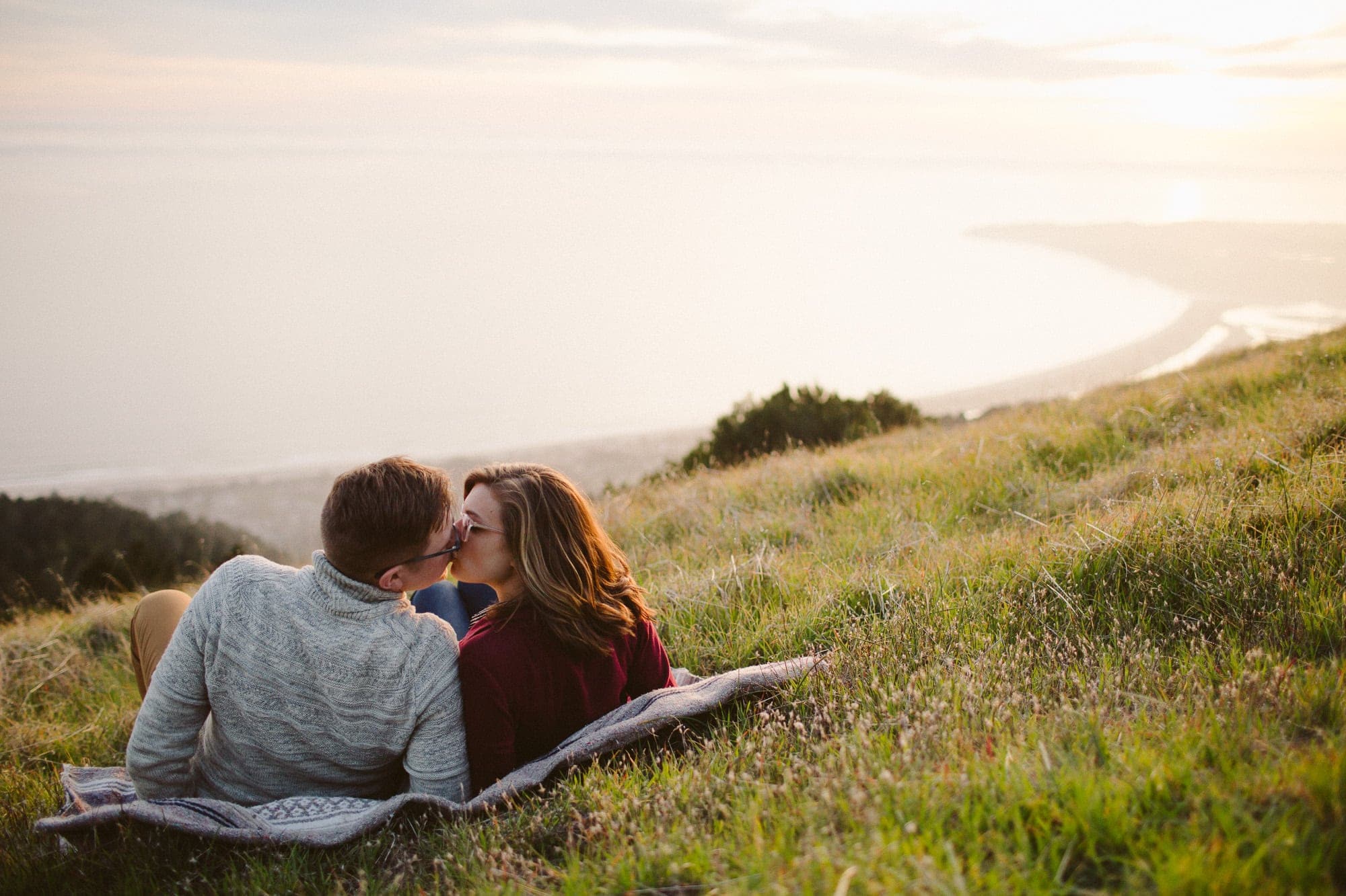 Couple kissing laying down on hill overlooking the ocean at sunset