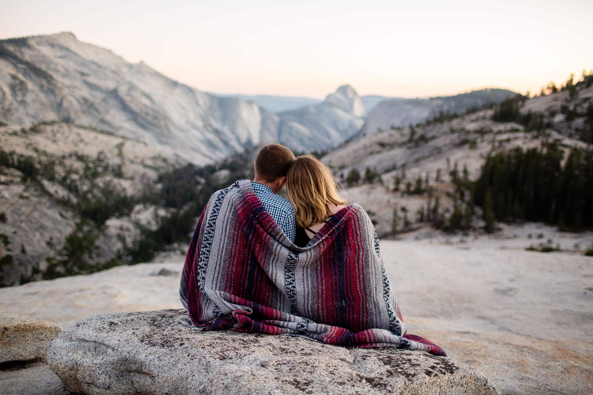 Man and woman wrapped in blanket sitting on mountain top looking at the view