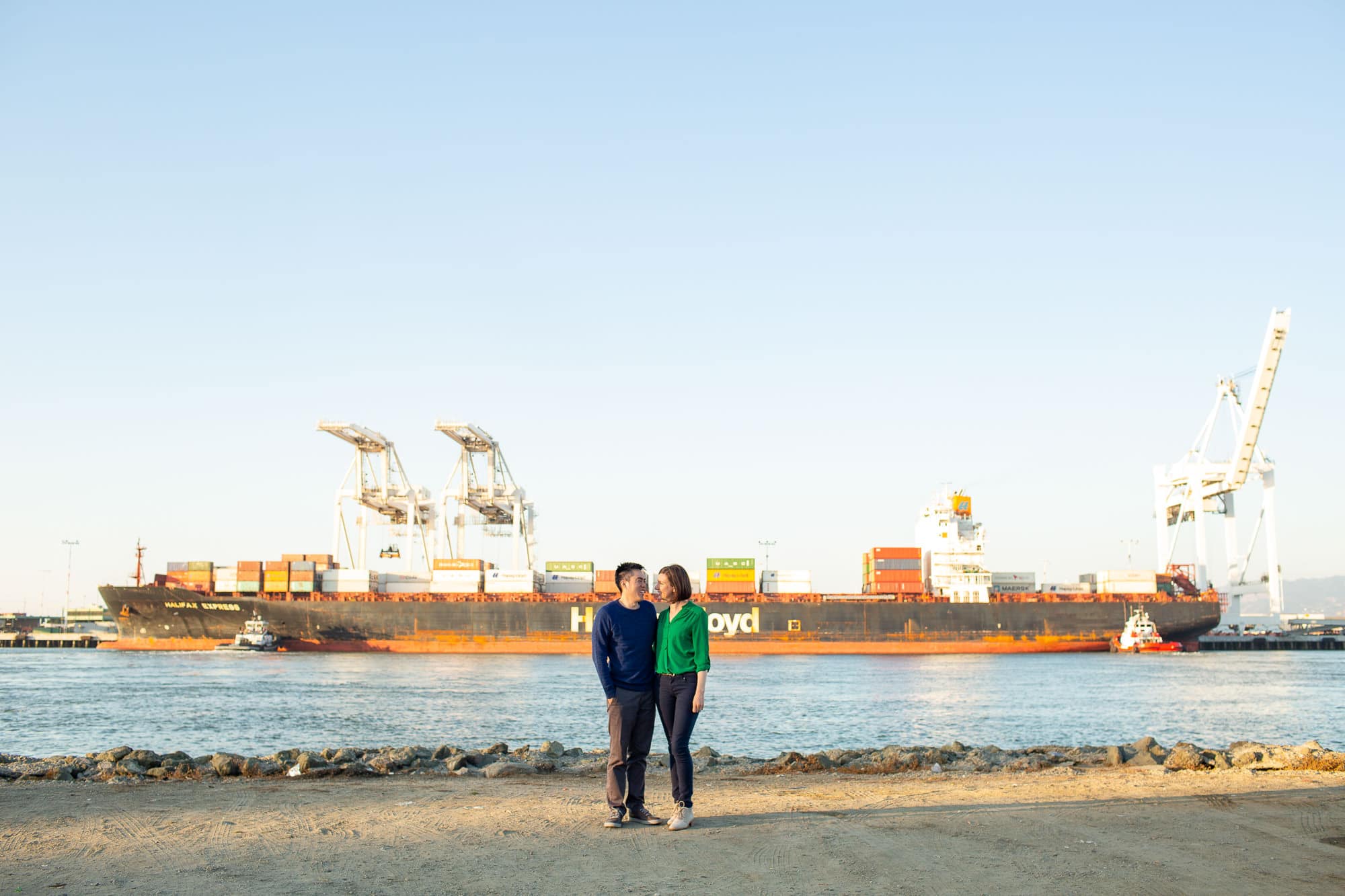 Couple standing looking at each other along the water with a cargo ship and shipping cranes in the background