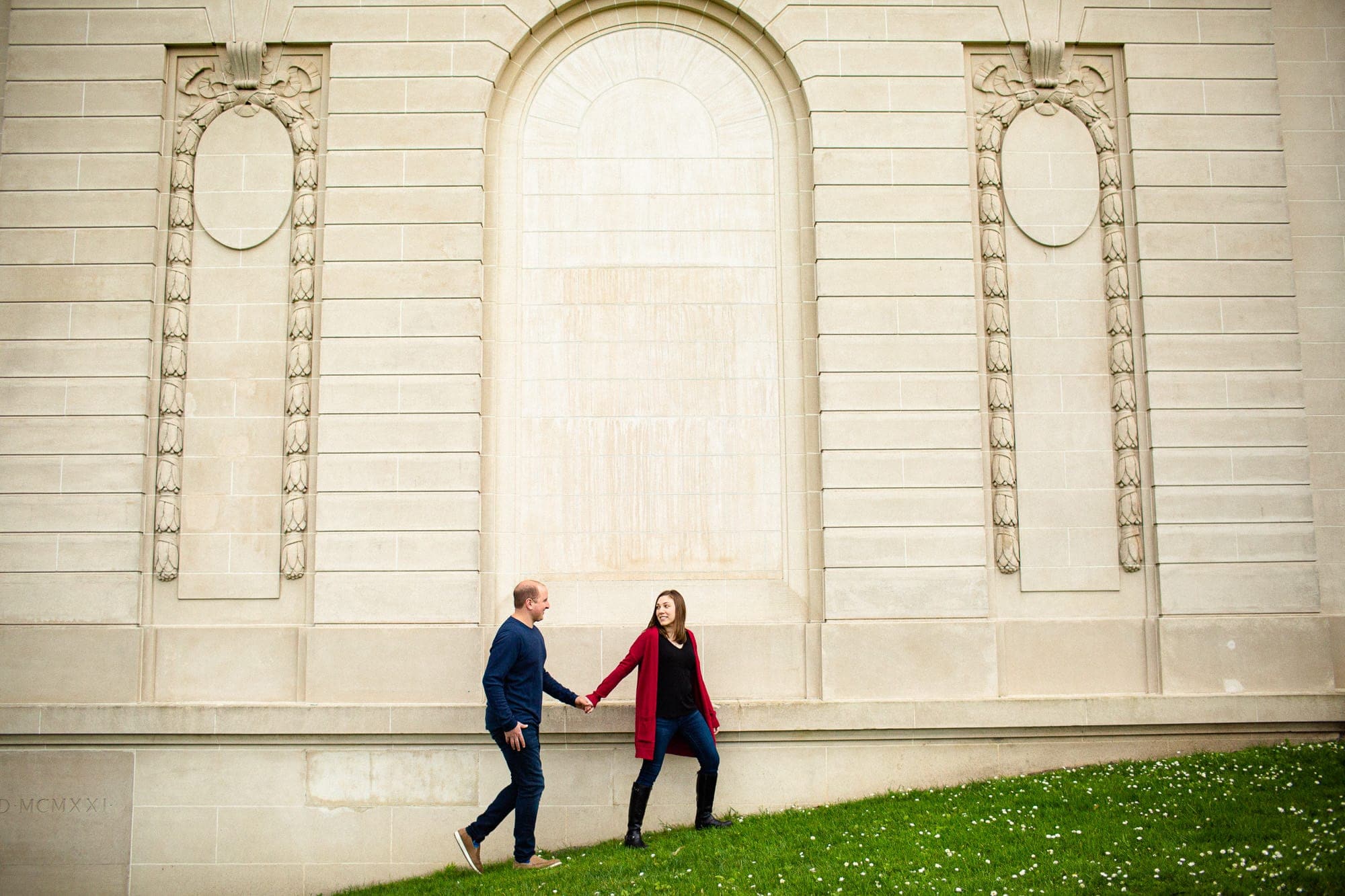 Woman leading man up a small hill next to a museum wall outside with green grass