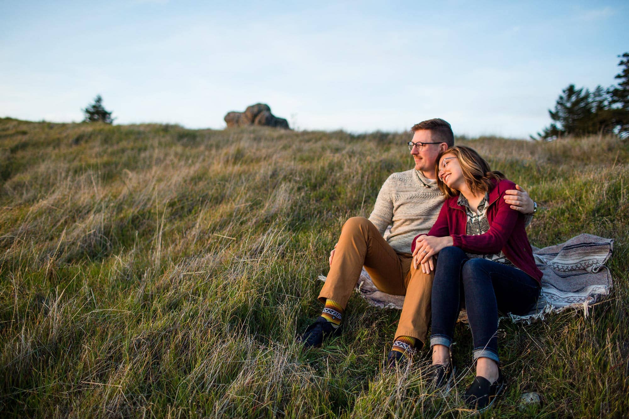 Couple sitting on blanket on grassy hill looking out at the sunset