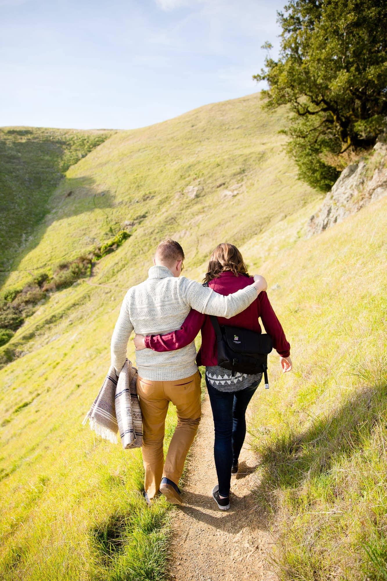 Couple with arms around each other facing away walking on a trail in the mountains with green grass