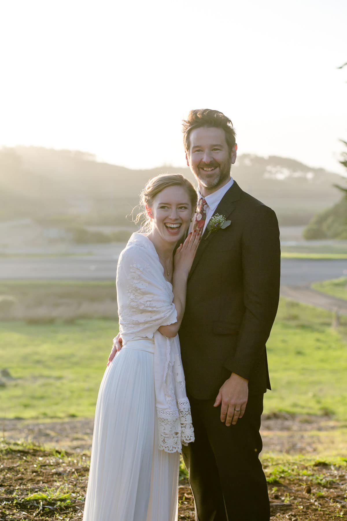 Bride and groom laughing and looking straight ahead outside on a grassy hill during sunset