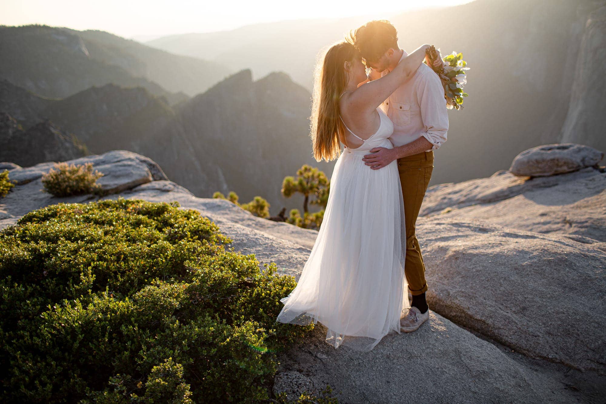 Woman in white dress and man in off white button up shirt and brown pants almost kissing on rocky area overlooking other mountains in Yosemite.