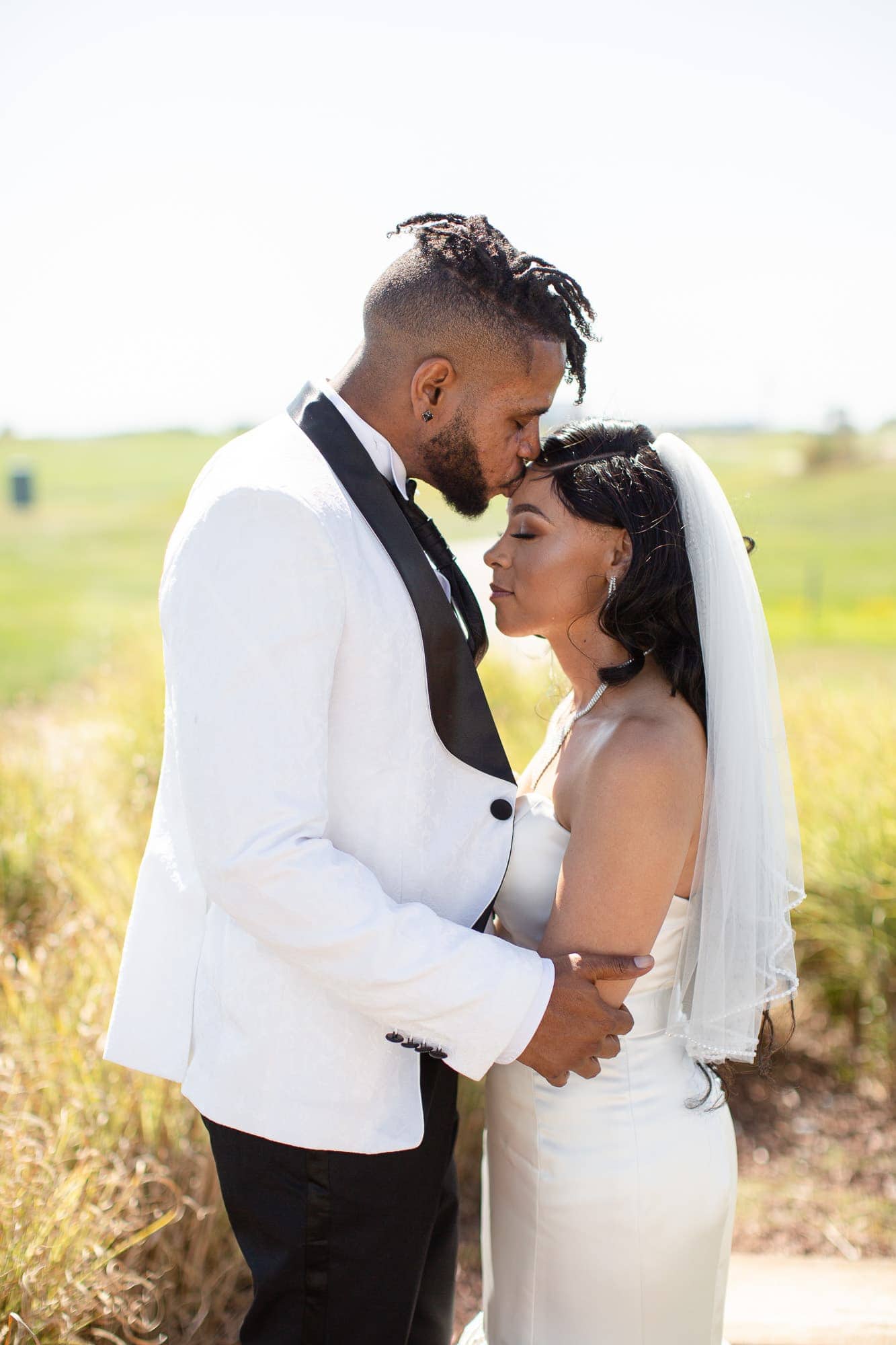 Groom in white tuxedo kissing bride on forehead outdoors