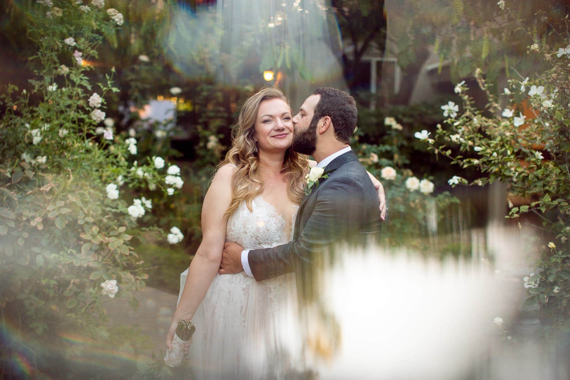 Groom kissing bride on cheek in a garden weeding in Northern California shot with a prism effect