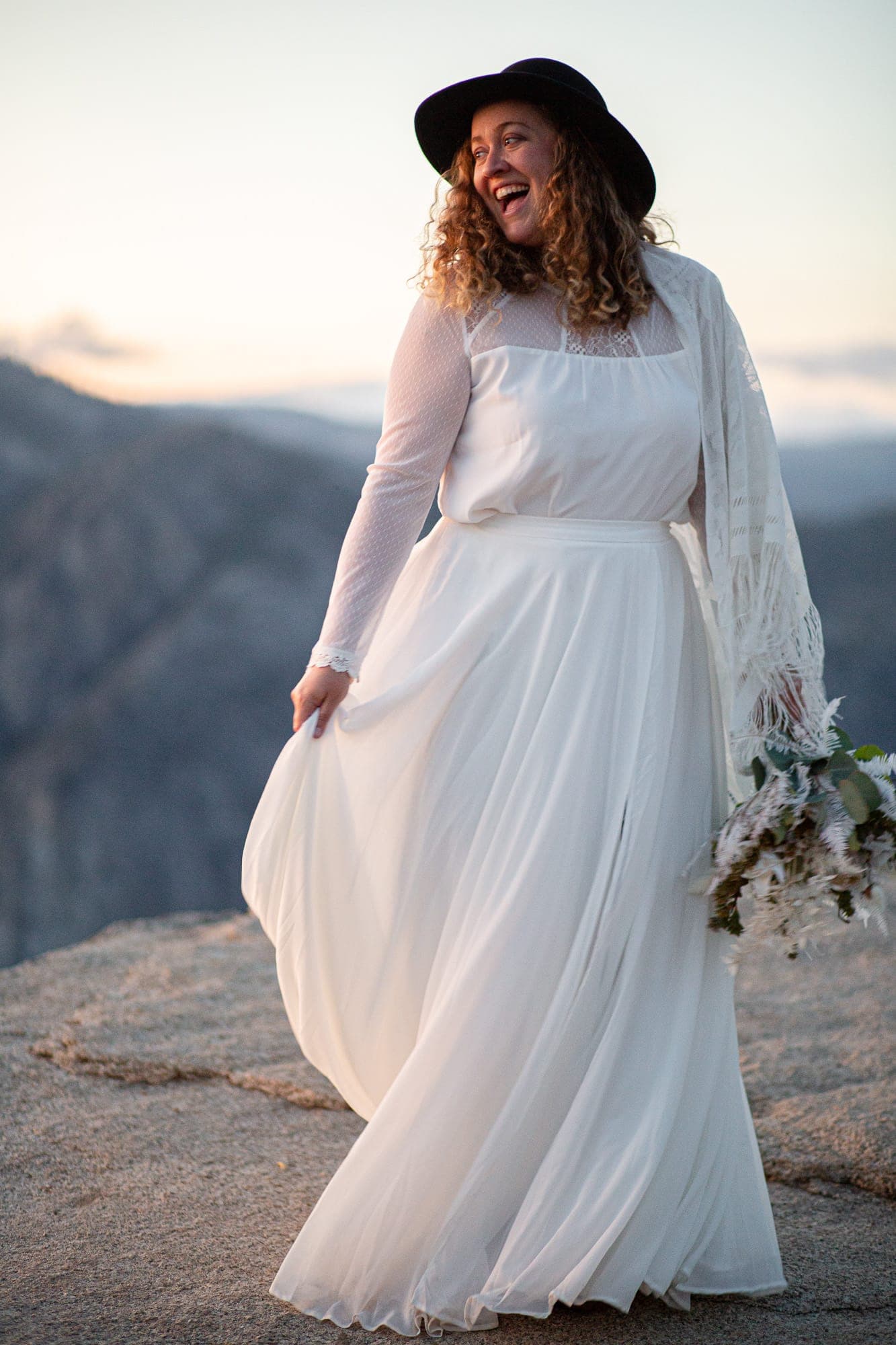 Boho bride in black hat and flowy dress posing in Yosemite at Taft Point at sunset