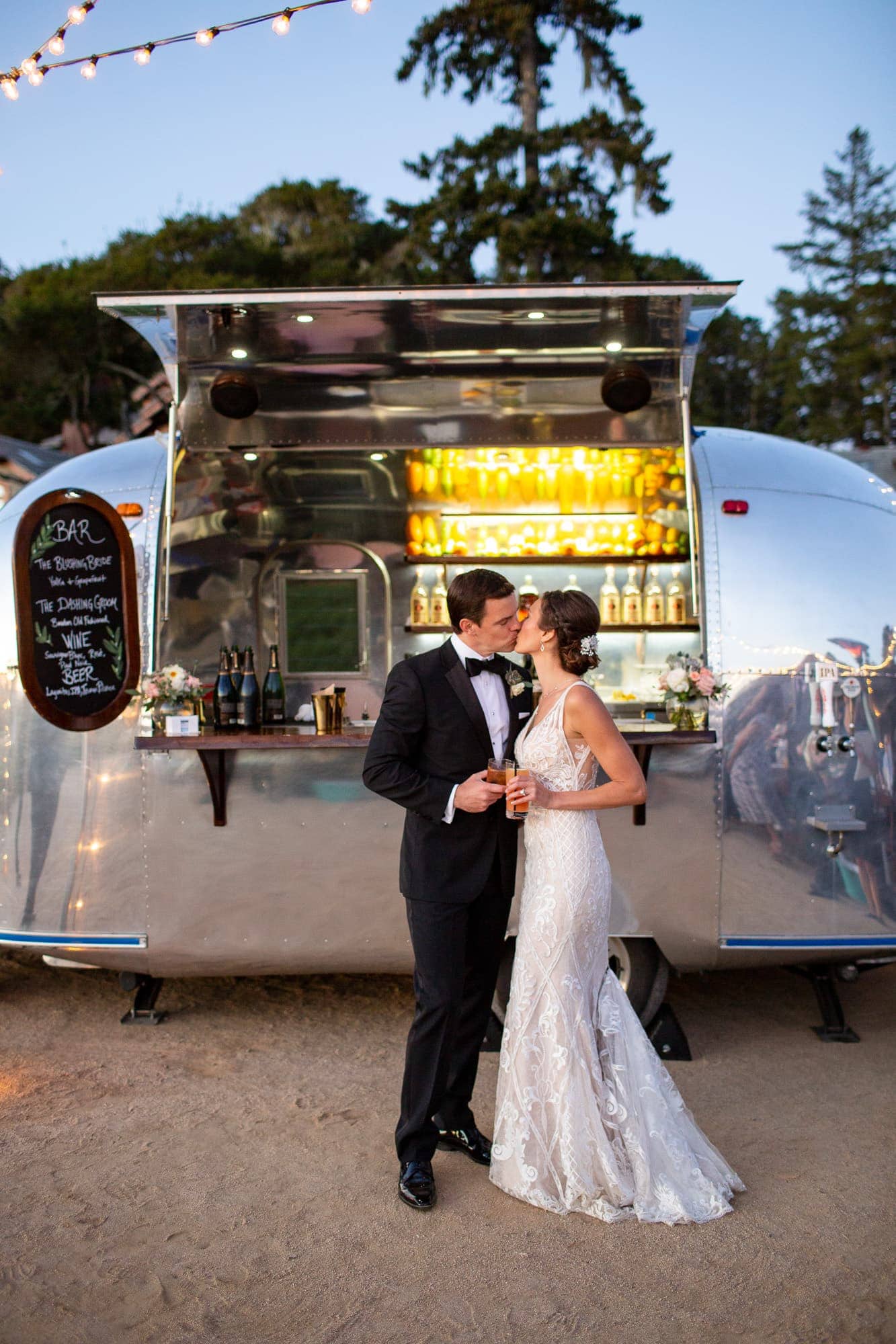 Groom in tuxedo and bride kissing and cheersing in front of an airstream bar at sunset
