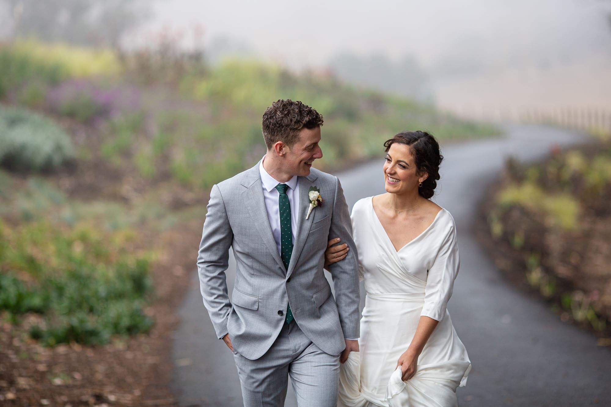 Groom and bride walk up winding road in foggy Tomales Bay Estate in grey suit and silk dress