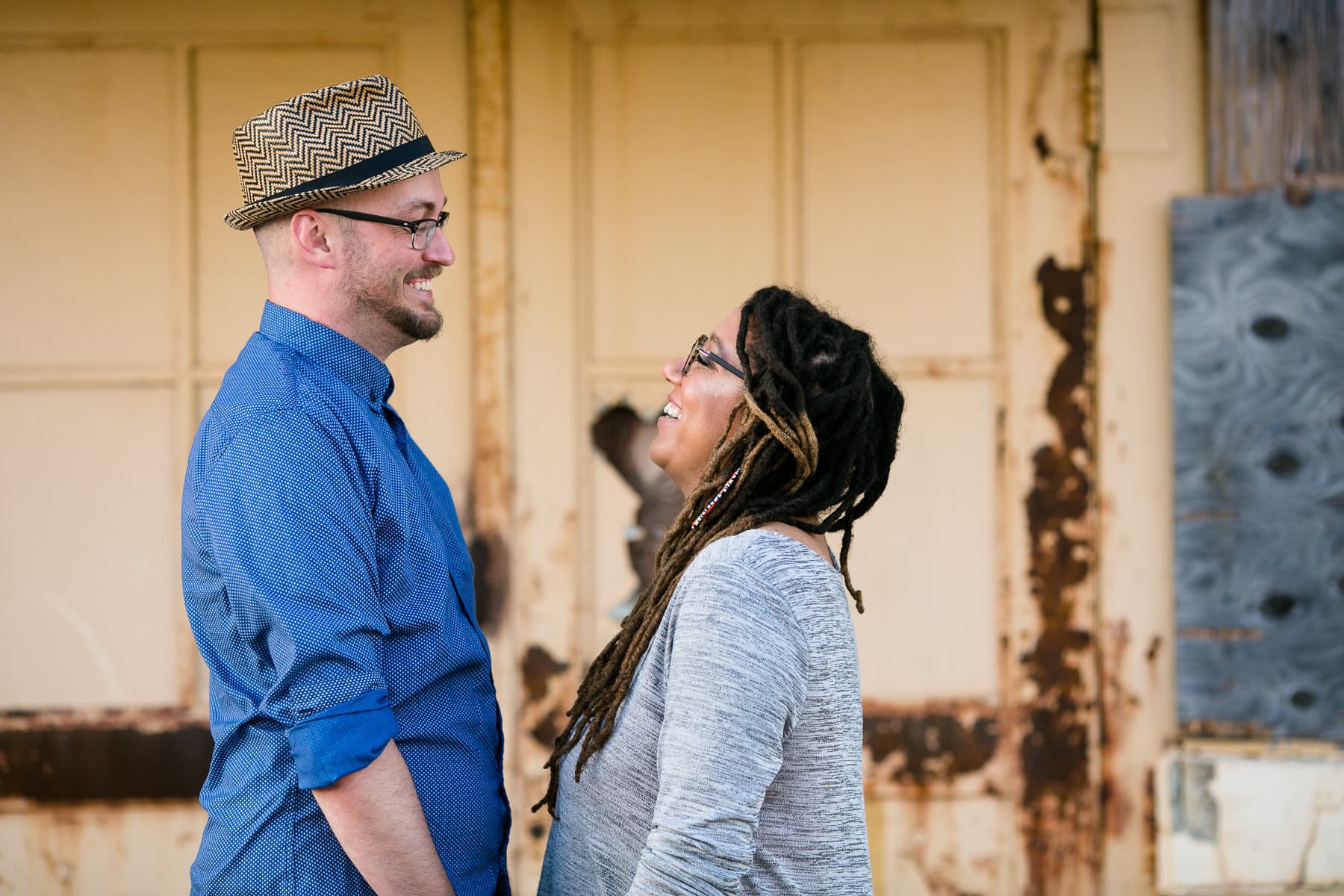Man in hat smiling and looking at woman with dreadlocks in front of rusted door