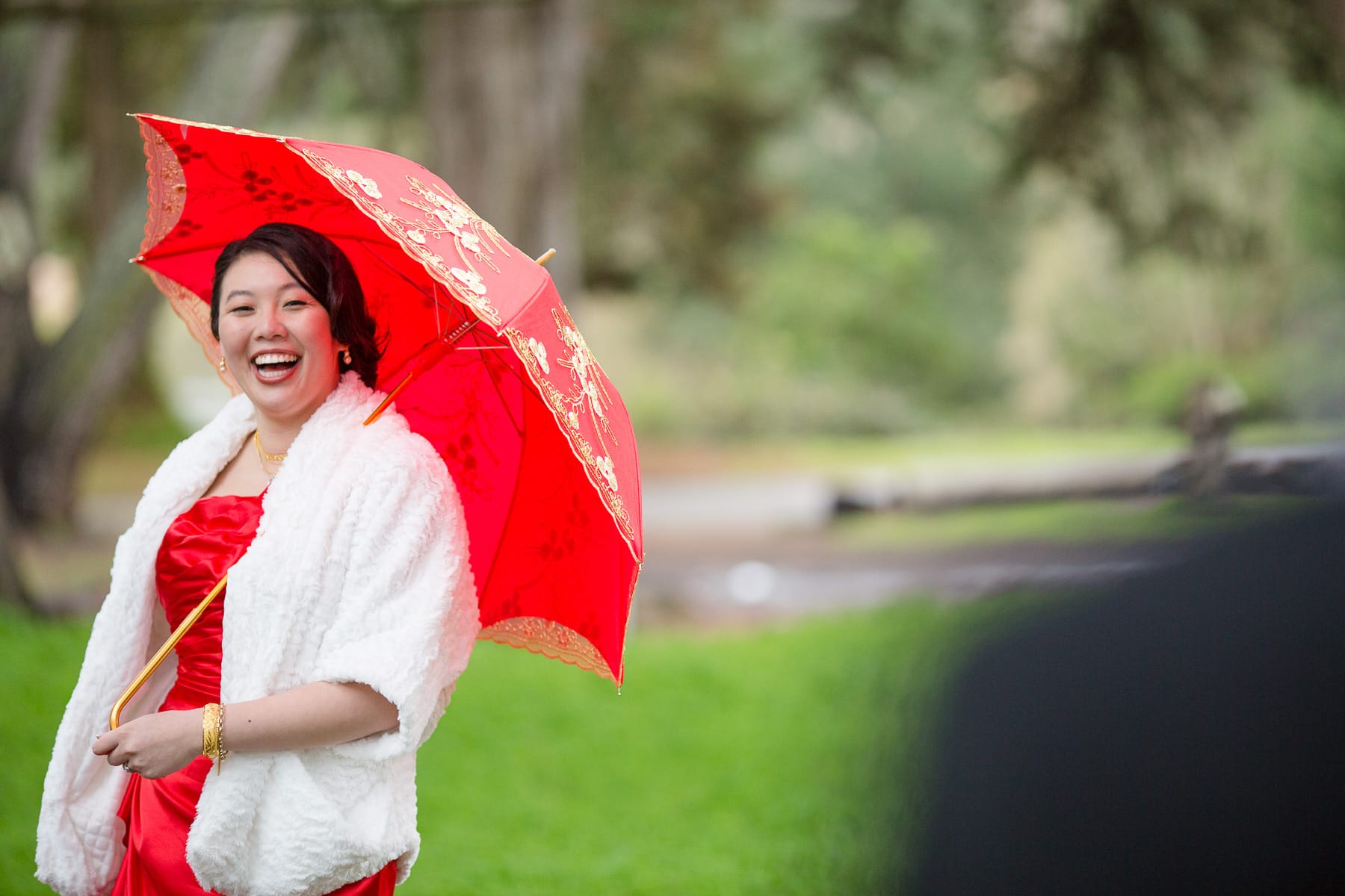 Bride in red dress and white shawl with red umbrella smiling while looking at her groom