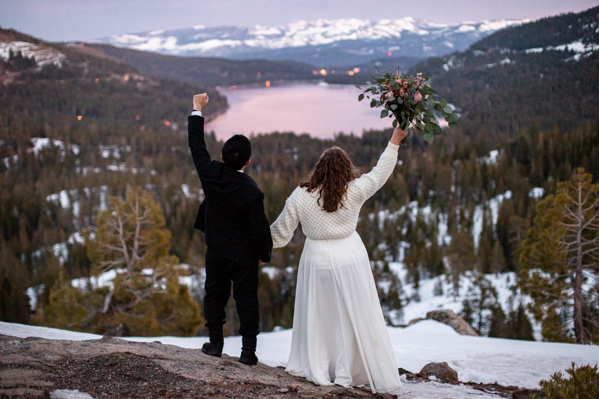 husband and wife standing in the mountains overlooking a lake down below at sunset celebrating with their hands in the air.