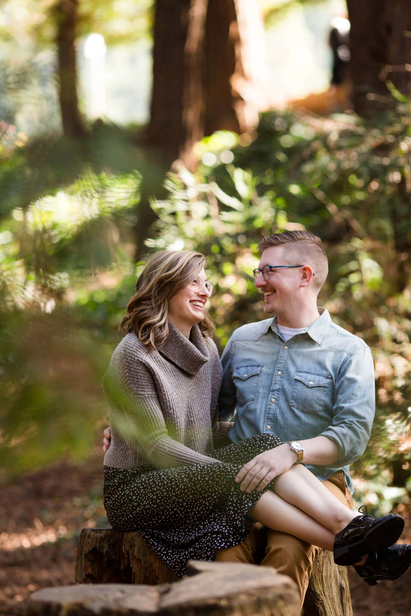 Couple sitting on a tree stump smiling and looking at each other in the woods