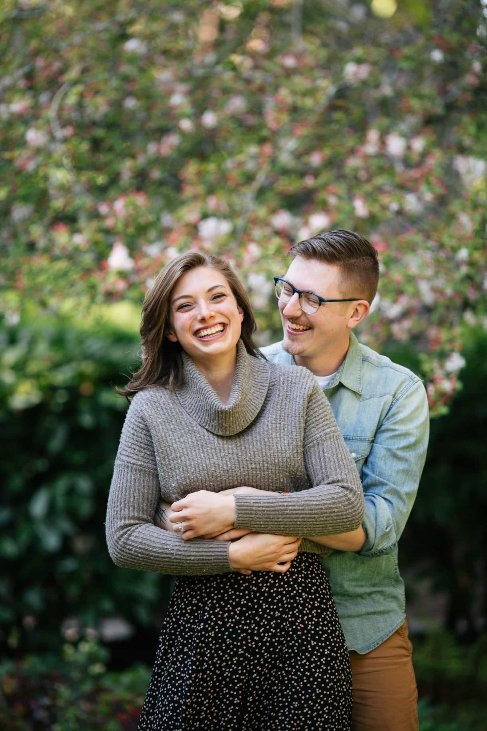 Man standing behind woman with arms around her laughing together in green floral garden