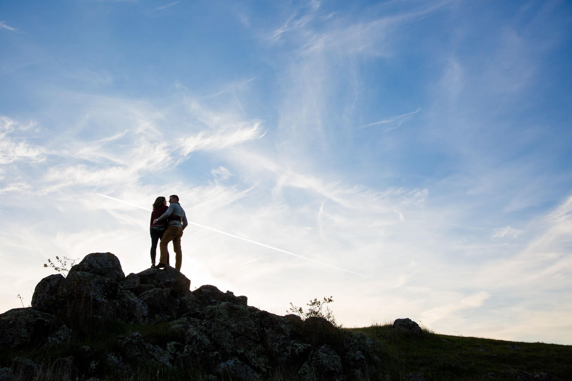 Silhouette of a couple standing on some tall rocks with clouds behind them