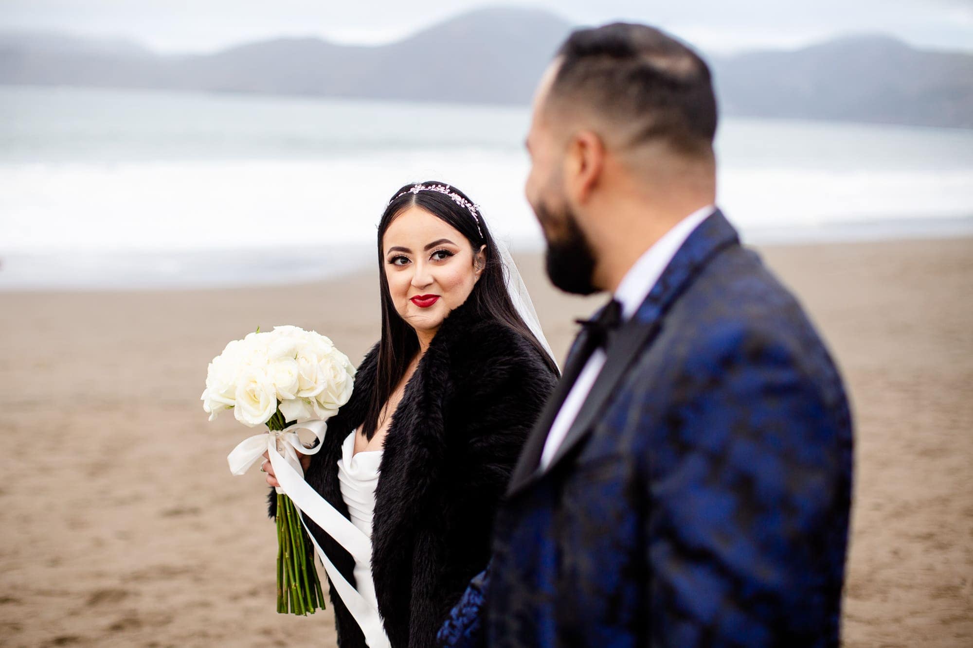 Bride looking at the groom while holding her bouquet and walking on the beach