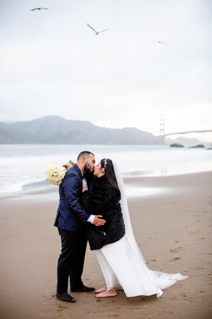 Husband and wife kissing on Baker Beach on new years with the Golden Gate Brideg in the background