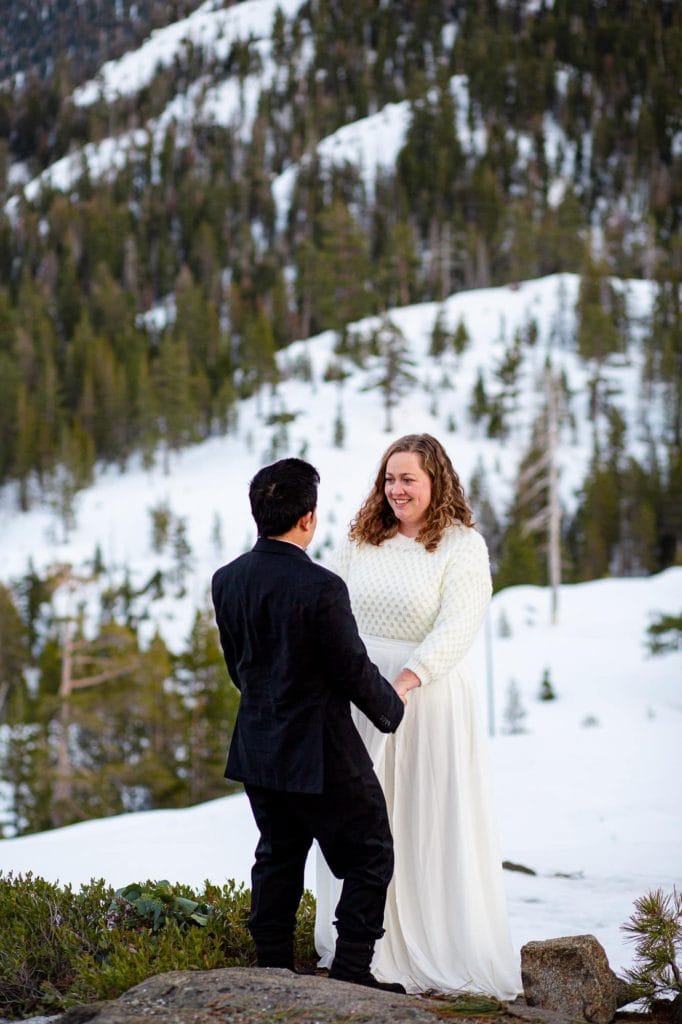 Groom with his back to the camera holding the brides hands who is facing the camera in a wintery snowy forest