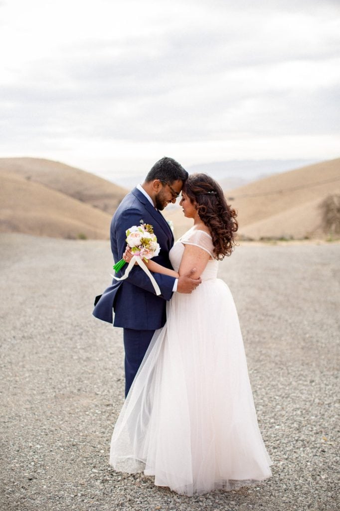 Groom and bride standing facing each other with their foreheads touching in front of rolling hills with yellow grass