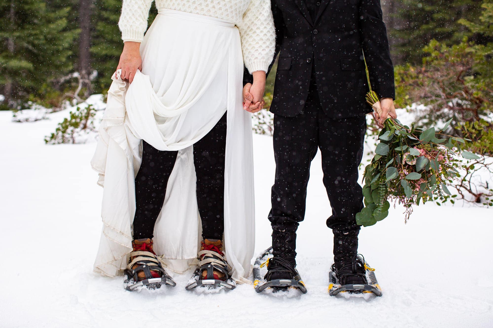 Bride and groom showing off their snowshoes in the snow with the groom holding the bouquet while the bride holds her skirt aside