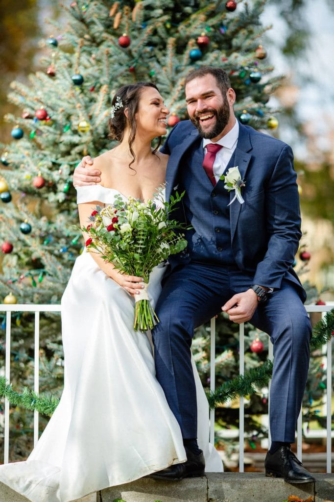 Couple in wedding clothes laughing and sitting on a white fence in front of a christmas tree