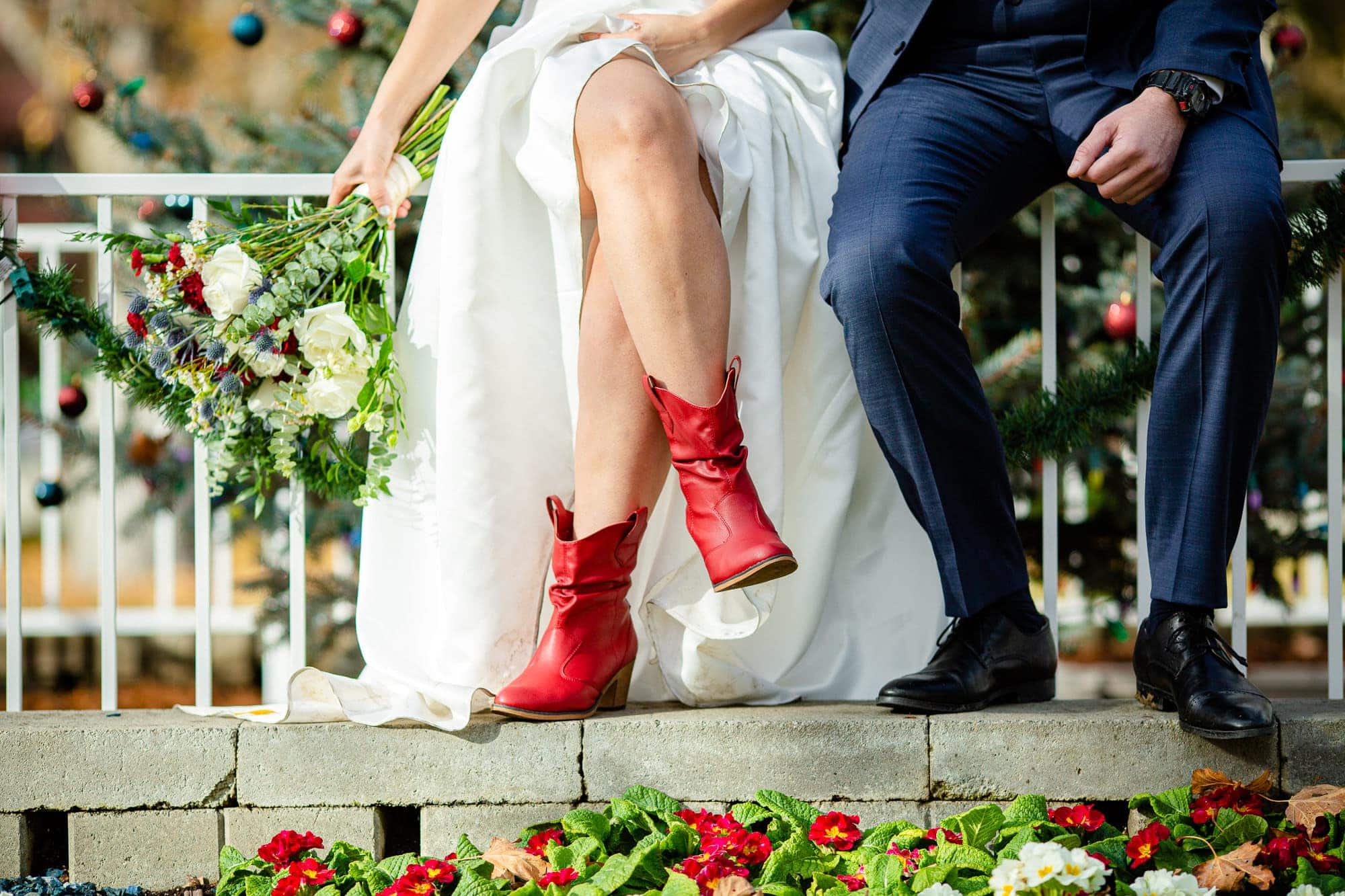 close up of a brides red cowboy boots as she sits on a fence in front of a christmas tree next to the groom