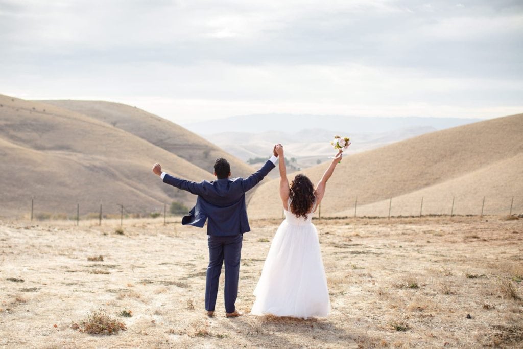 husband and wife with backs to the viewer celebrating in wedding attire with arms in the air on golden grassy hills